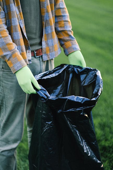 Close-up of a person wearing a checked yellow and grey jacket, green gloves, and grey trousers, standing outdoors on a grassy area, holding open a large black plastic rubbish bag lined with a grey inn